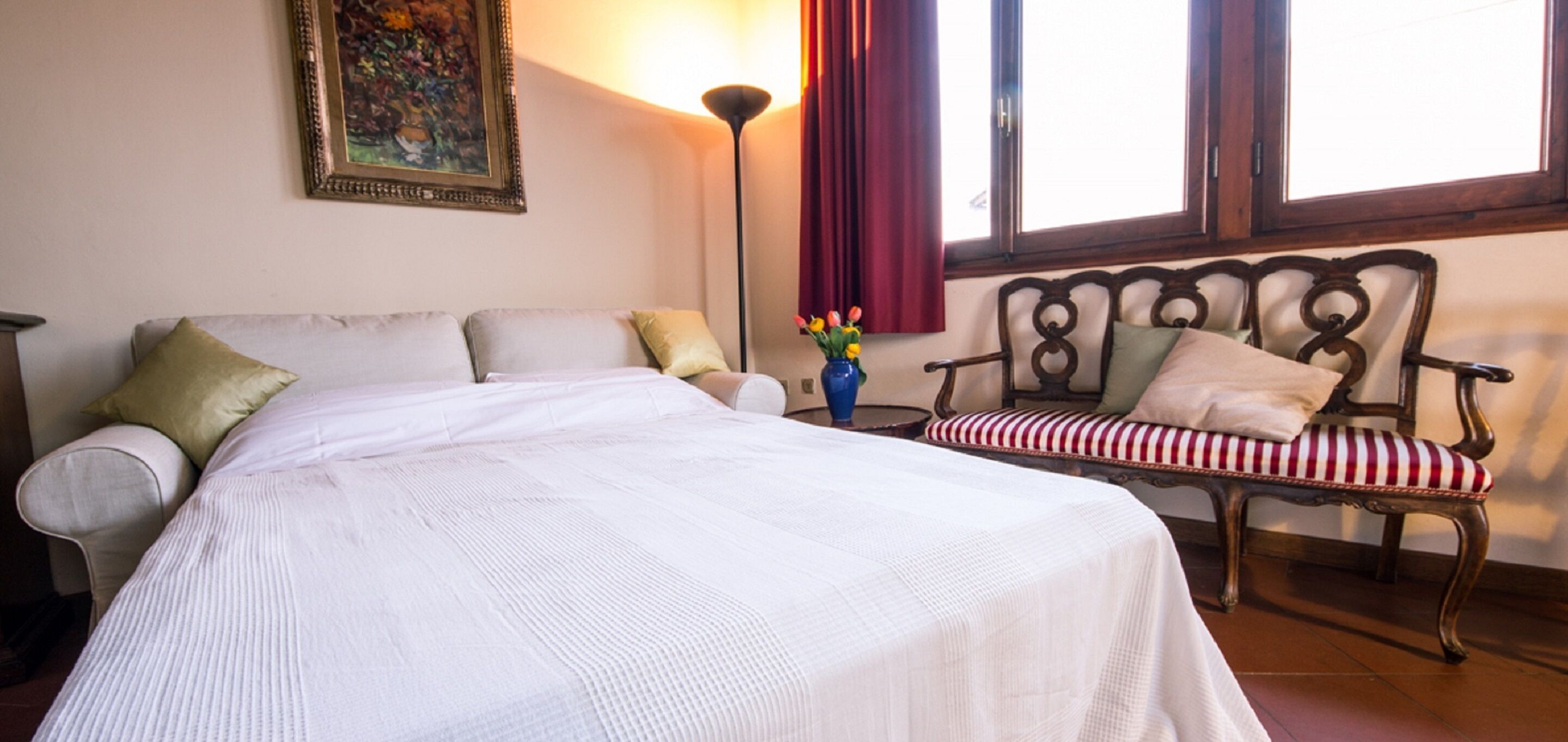 Une chambre avec un grand lit, une chaise dotée d’un coussin à rayures rouges et blanches, un vase rempli de fleurs et un tableau accroché au mur.