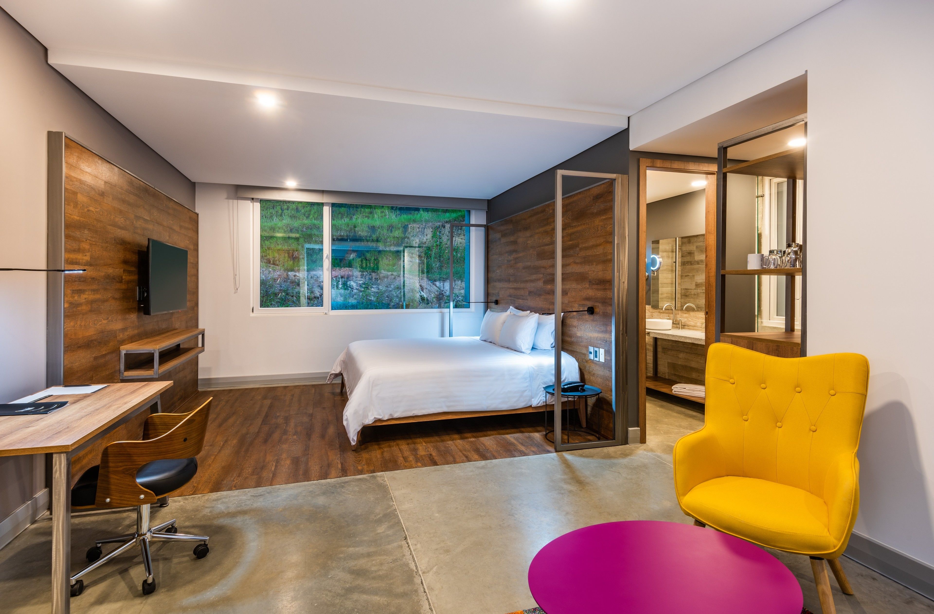 A modern hotel room with a large bed, a desk, a chair, and a bright yellow armchair.