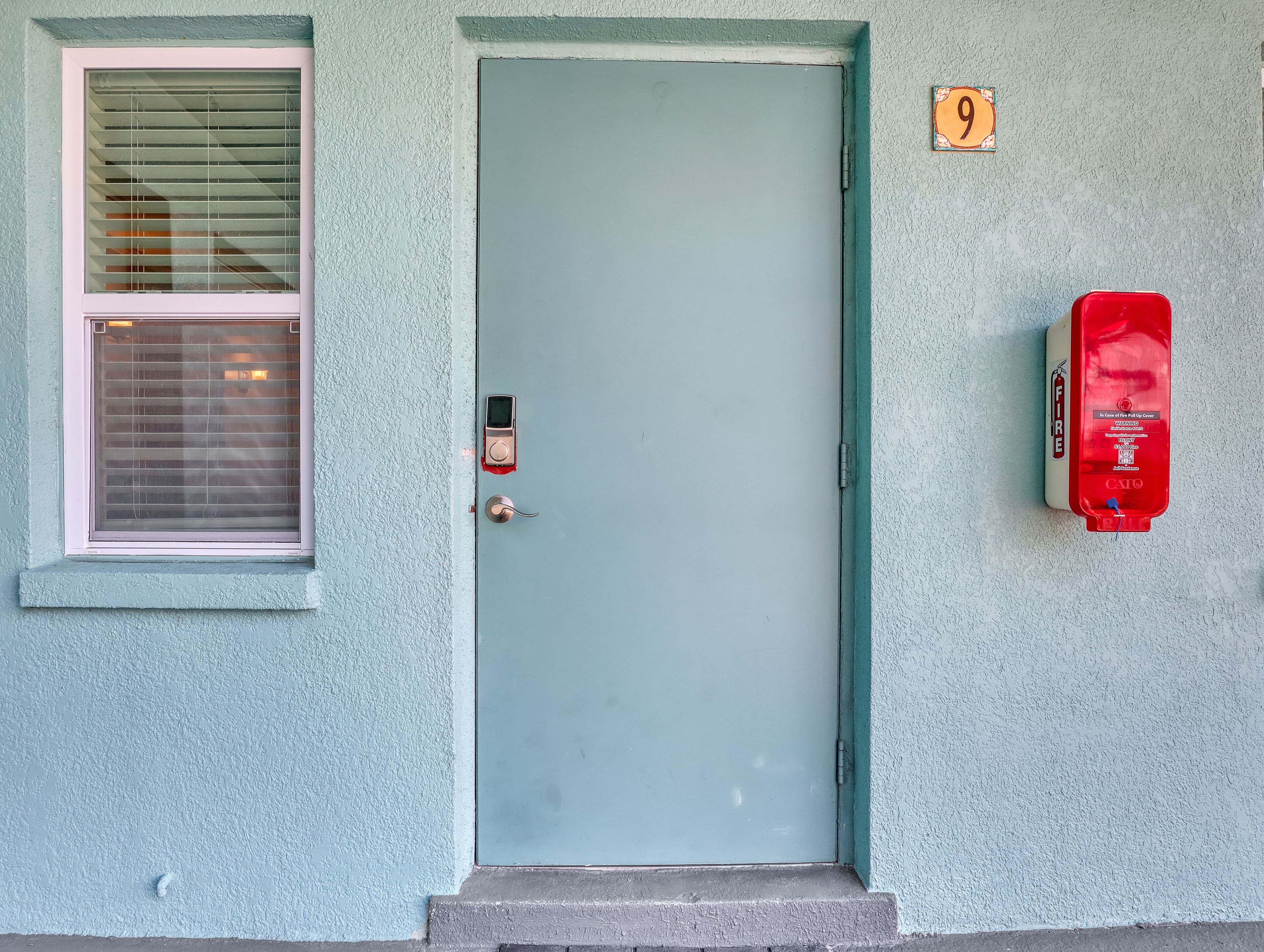 Une porte avec une plaque numérotée, une fenêtre avec des stores et un extincteur fixé au mur.
