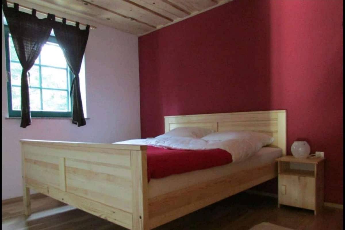 A single bed with a wooden frame and white bedding, a bedside table with a lamp, and a window with curtains.