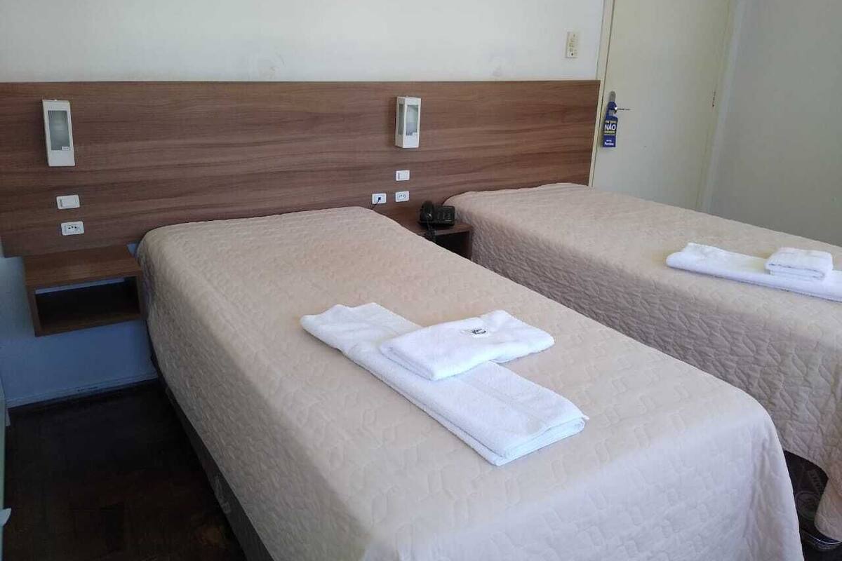 Two single beds with beige bedding, a wooden headboard, and a bedside table with a phone and a bottle of hand sanitizer.