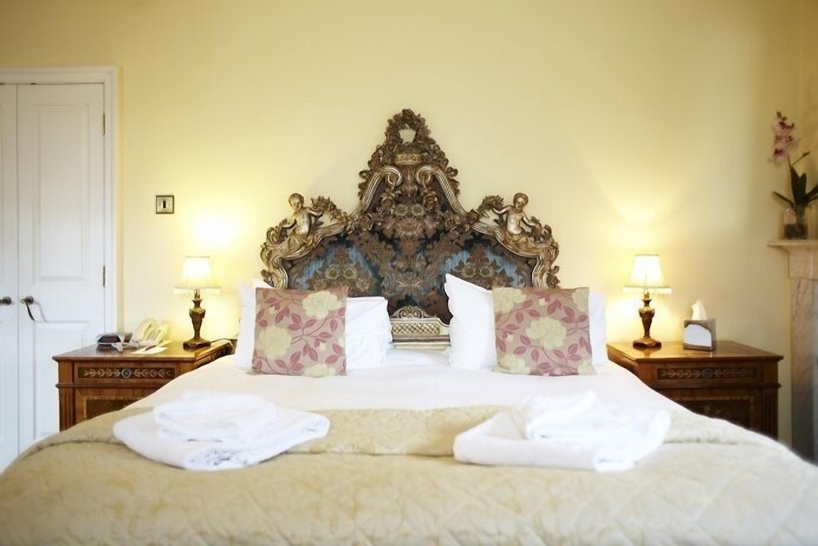 A hotel room with a large bed, ornate headboard, floral pillows, bedside lamps, and a telephone on a wooden nightstand.
