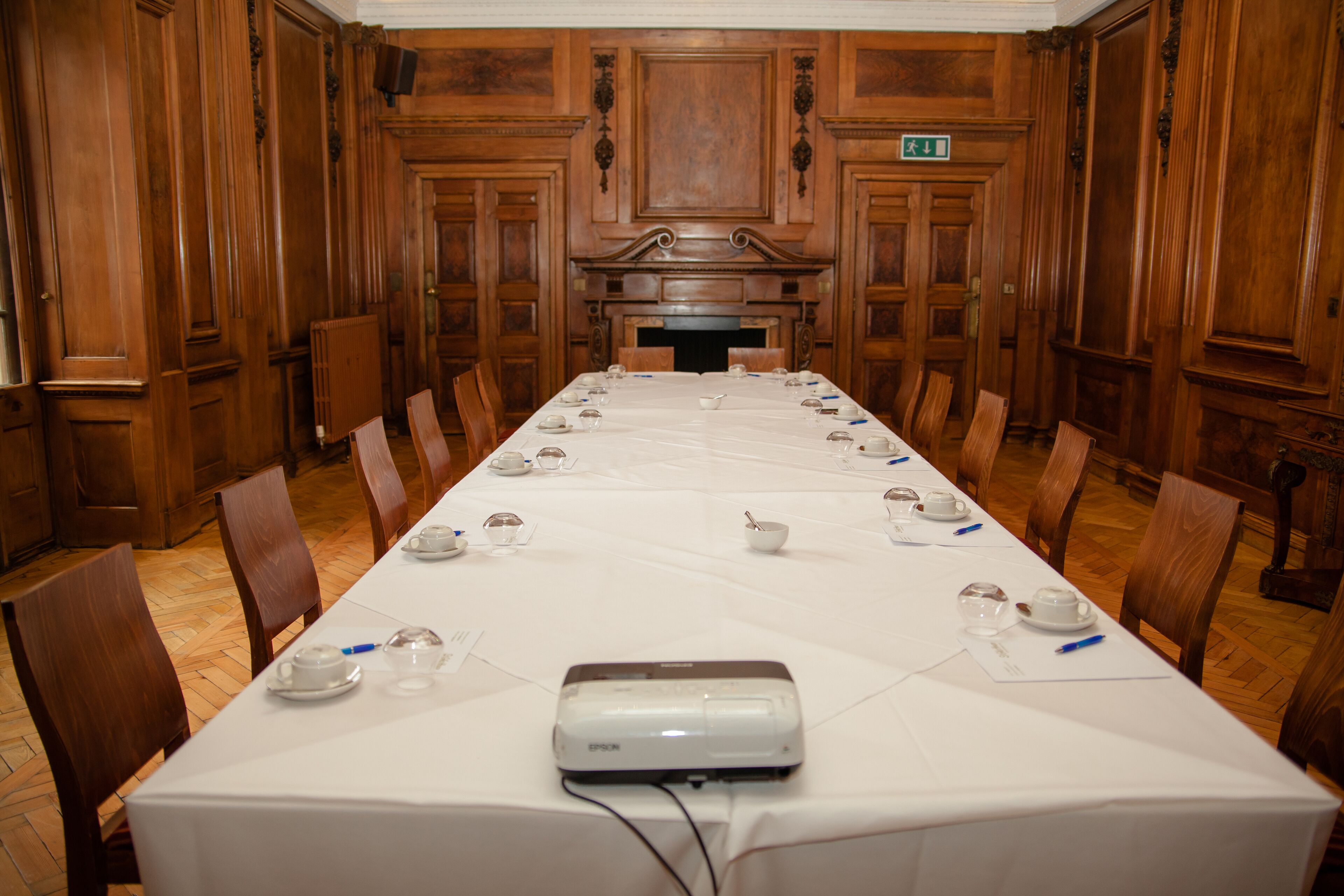 A conference room with a long table set for a meeting, equipped with a projector, and surrounded by wooden paneling and chairs.