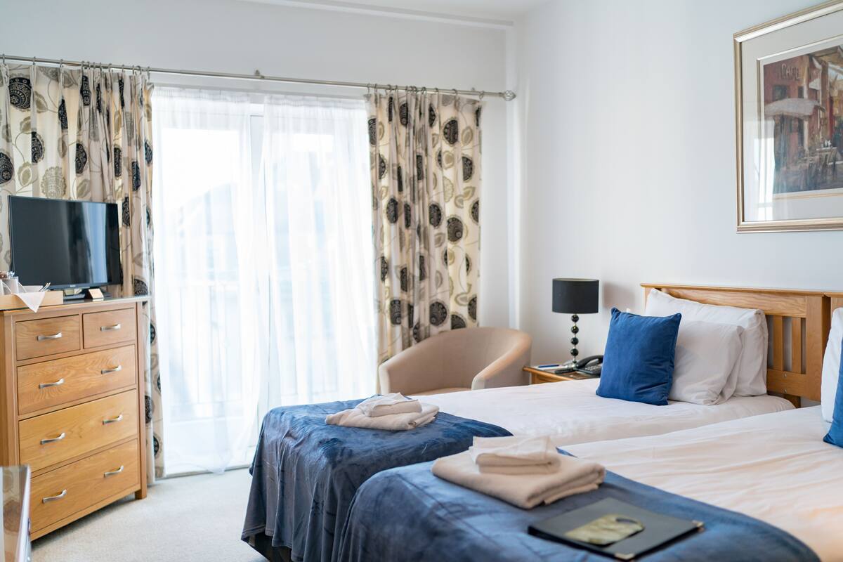 A hotel room with a bed, a chair, a desk with a lamp, a TV on a dresser, and a window with curtains.