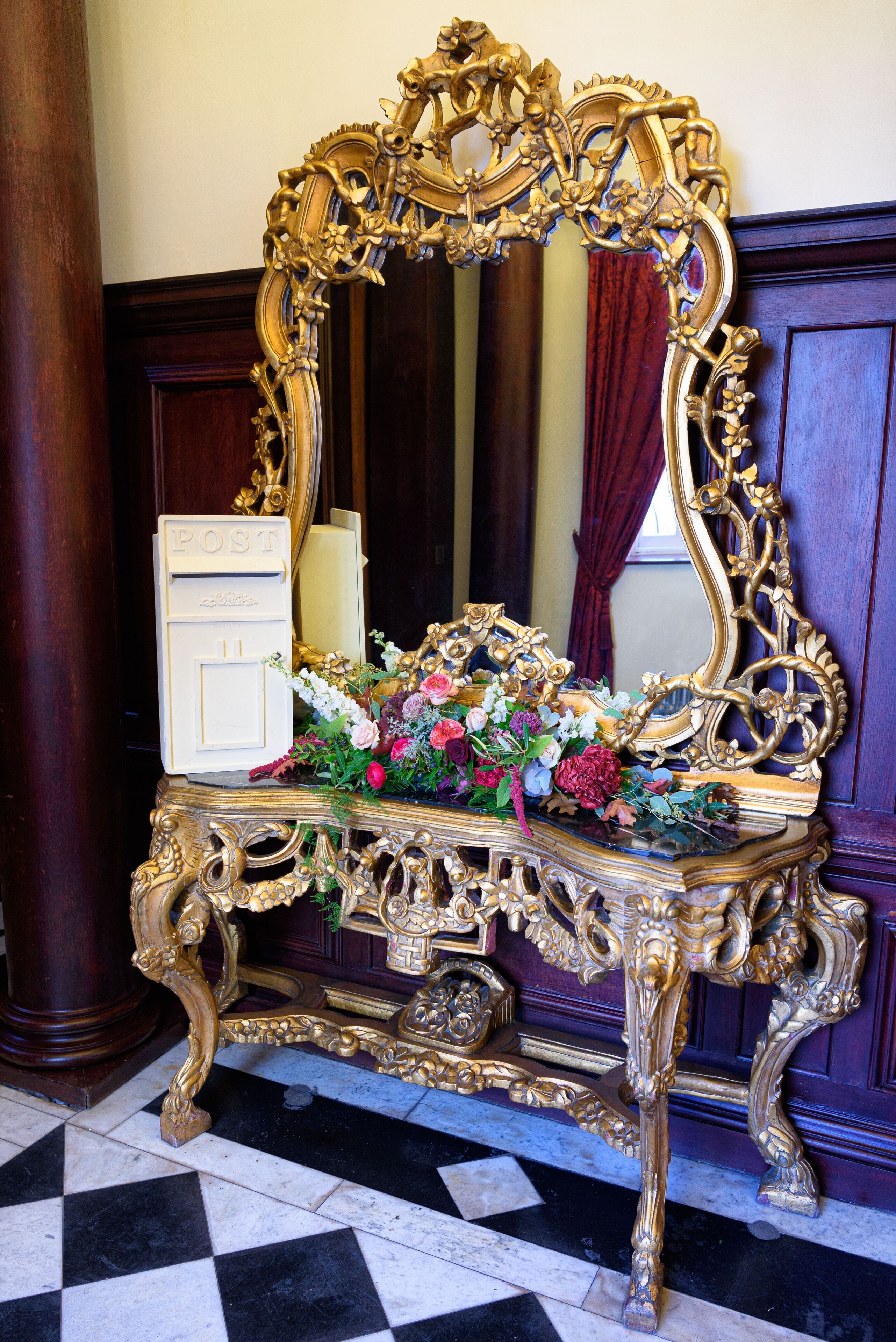 An ornate gold-framed mirror with a floral arrangement on a marble floor.