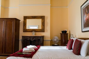 A neatly made bed with a red throw blanket, a wooden wardrobe, a fireplace, a wall-mounted mirror, and a fan.