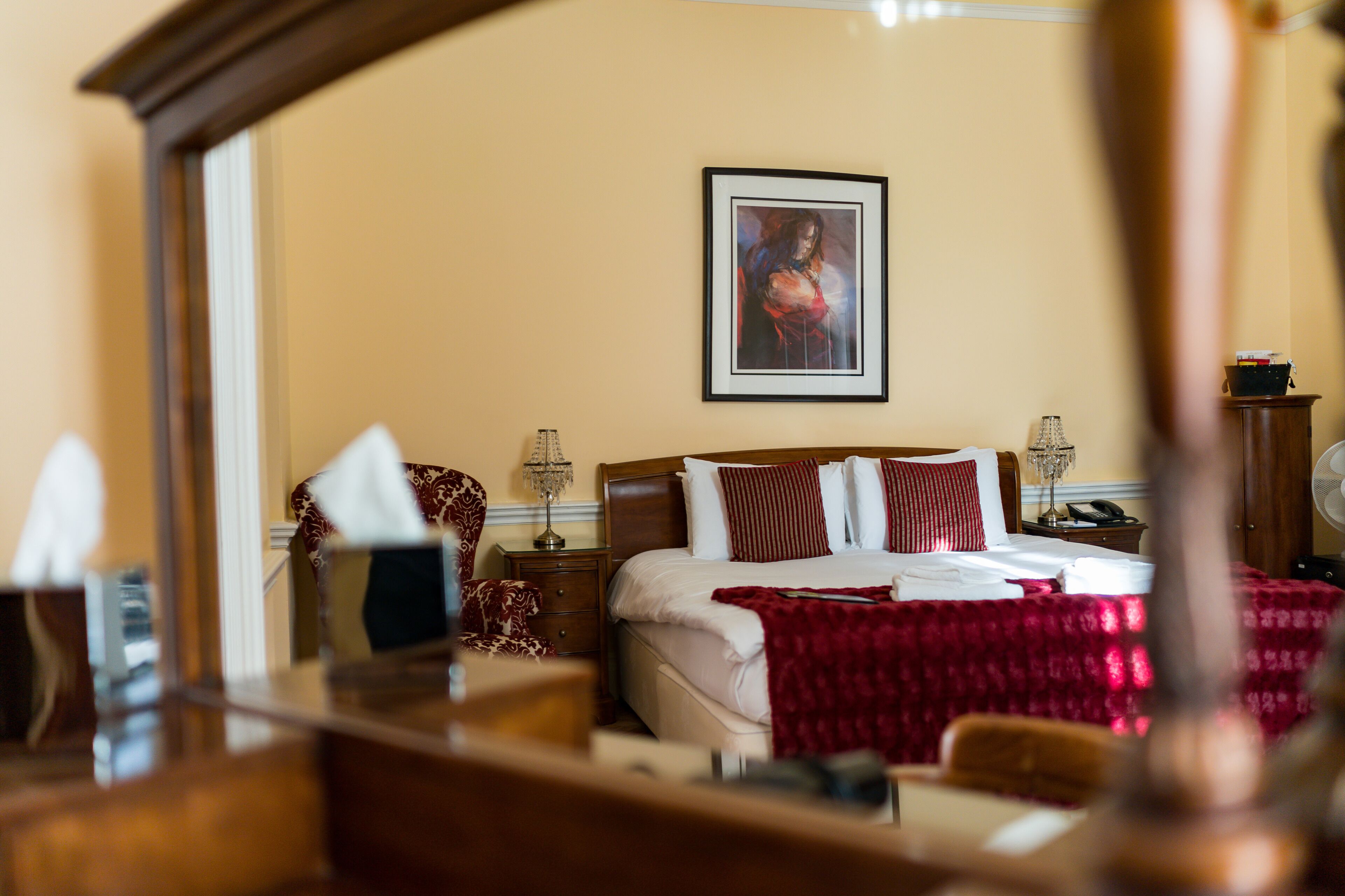 A hotel room with a large bed, a bedside table with a lamp, a framed picture on the wall, and a mirror reflecting part of the room.