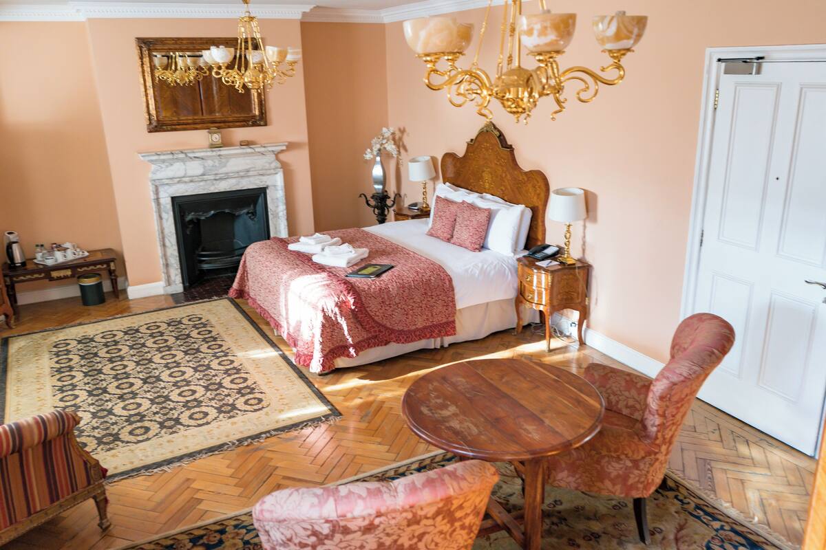 A bedroom with a large bed, a fireplace, a chandelier, a round wooden table, and two upholstered chairs.
