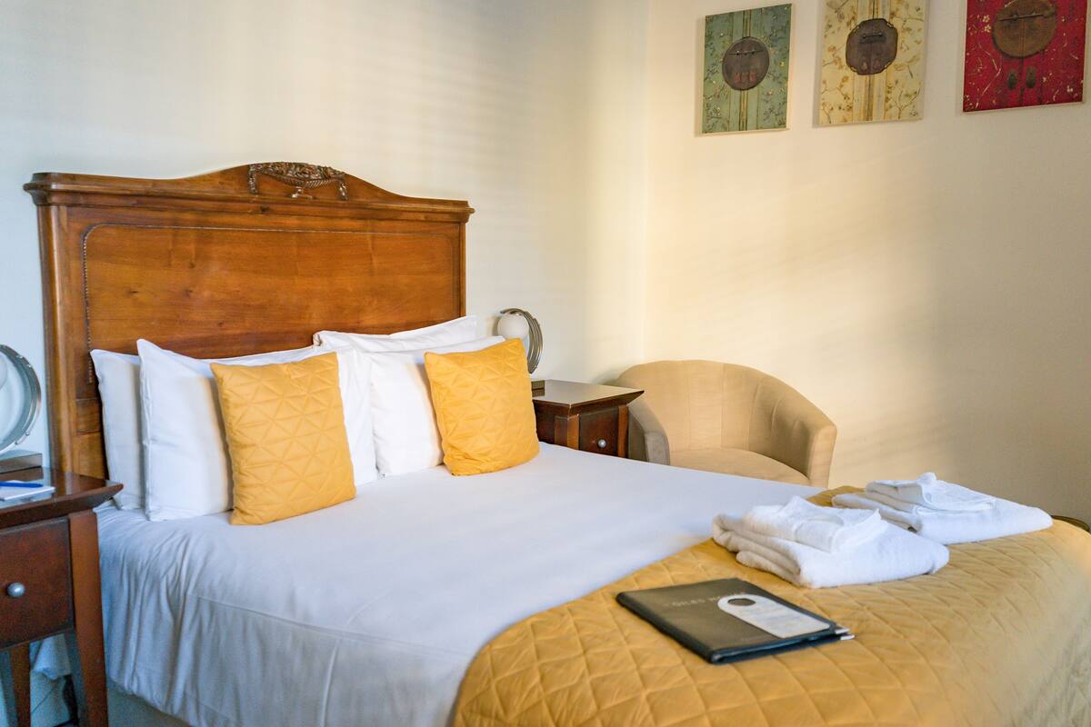 A neatly made bed with a wooden headboard, white and yellow pillows, and a laptop on the bed. There is a chair and a small table beside the bed.