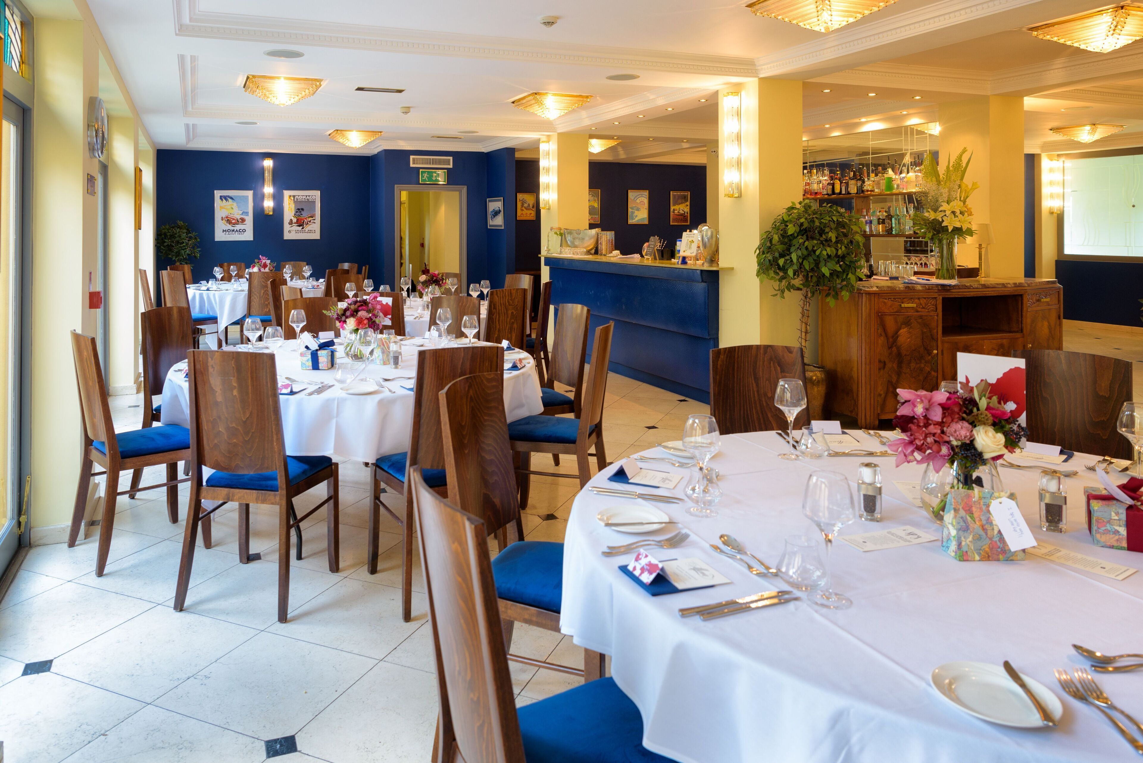Un restaurante con mesas redondas dispuestas para comer, sillas azules y una zona de bar con una planta y utensilios.