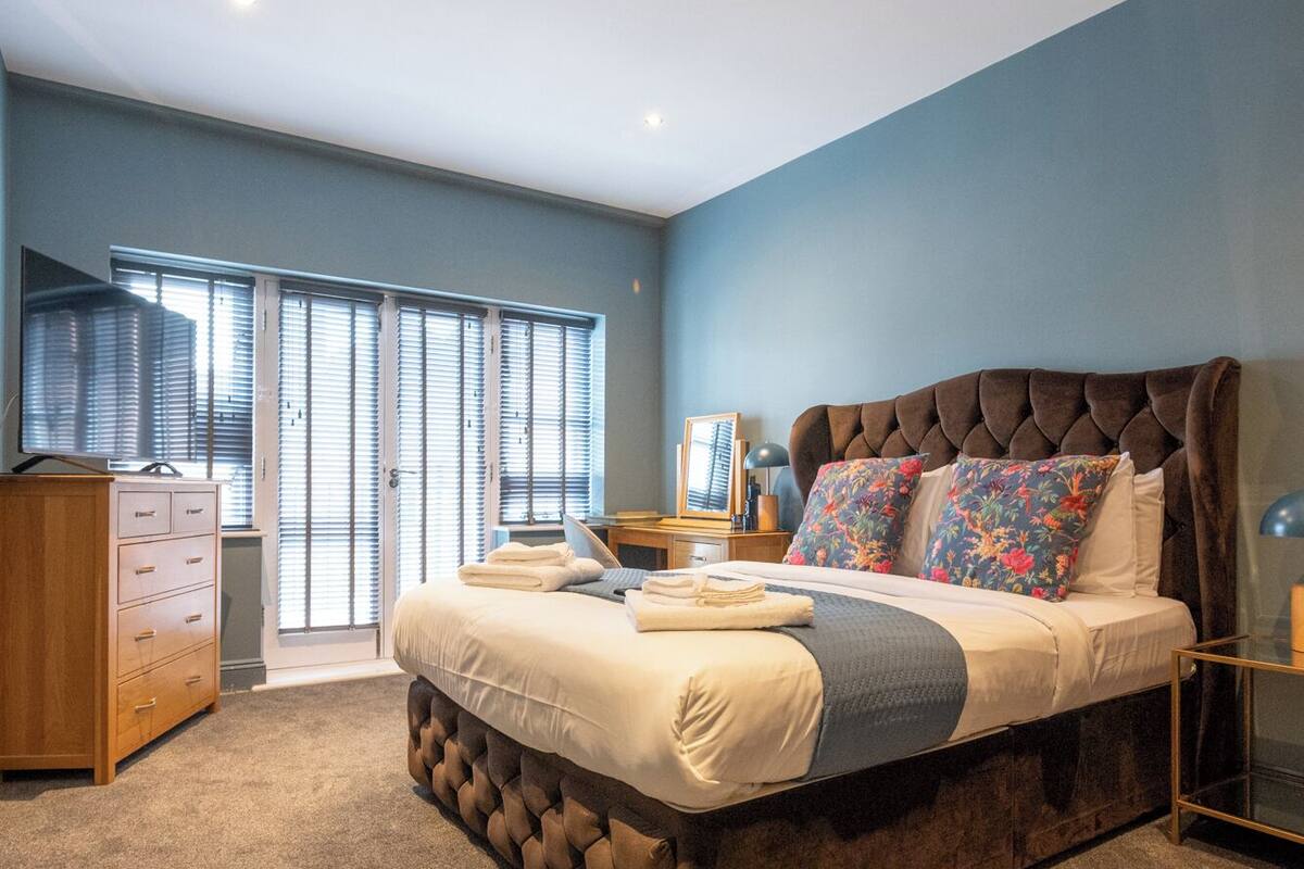 A bedroom with a large bed, a wooden dresser, and a view of the outside through a window with blinds.