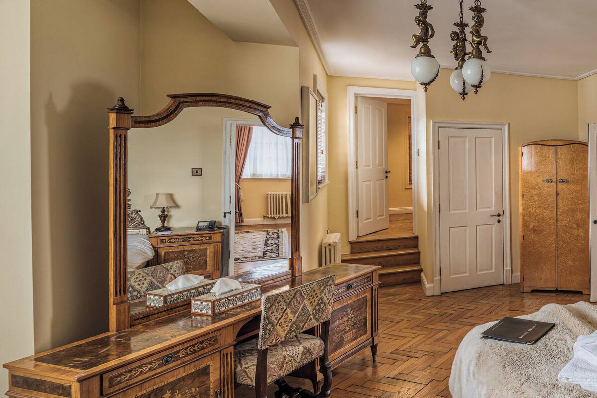 A hotel room with a large bed, a wooden dresser, a desk with a chair, a mirror, and a wardrobe.