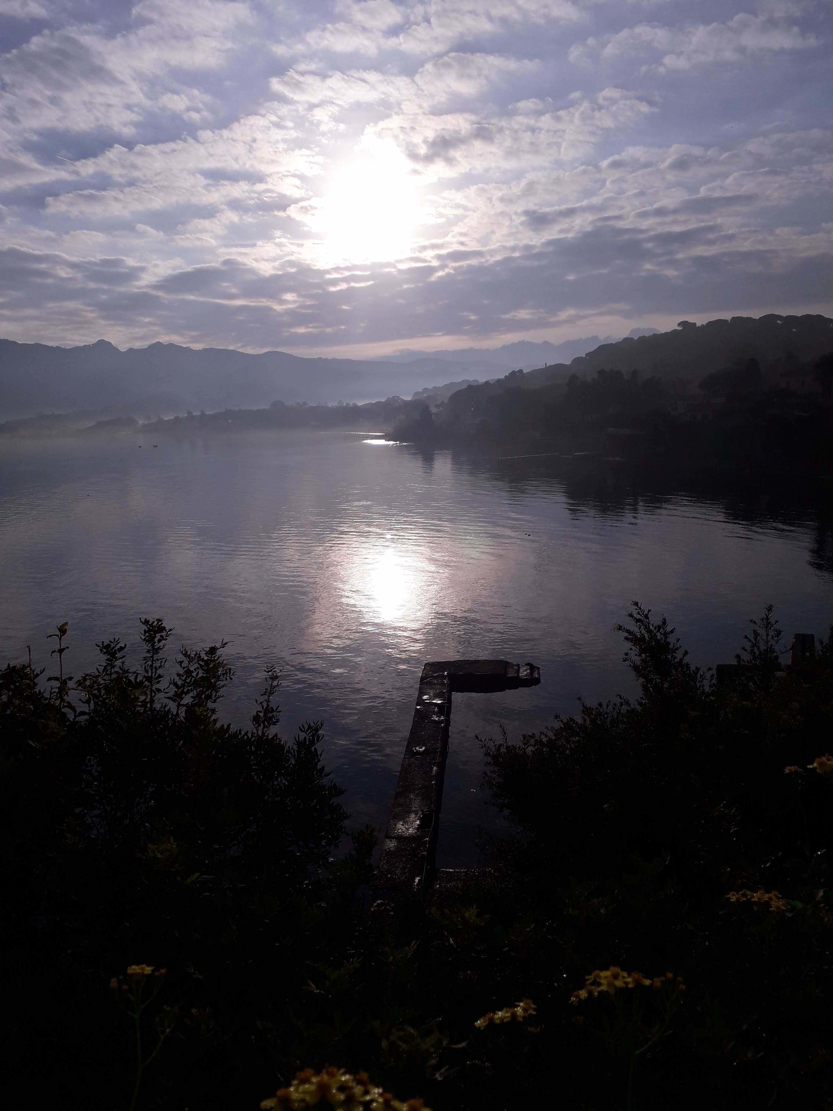 A calm lake with a wooden dock extending into the water, surrounded by vegetation and distant mountains under a partly cloudy sky.