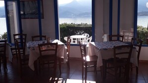 A dining area with tables and chairs, a chandelier, and a view of a mountain and lake. - Hotel Grotte del Paradiso (Portoferraio)