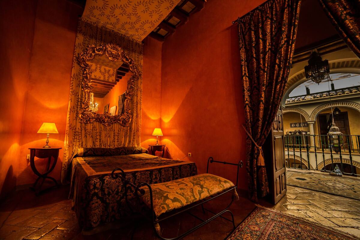 Une chambre à coucher à l’éclairage chaleureux, avec un lit, deux tables de chevet dotées de lampes, un banc et une vue sur une cour extérieure.