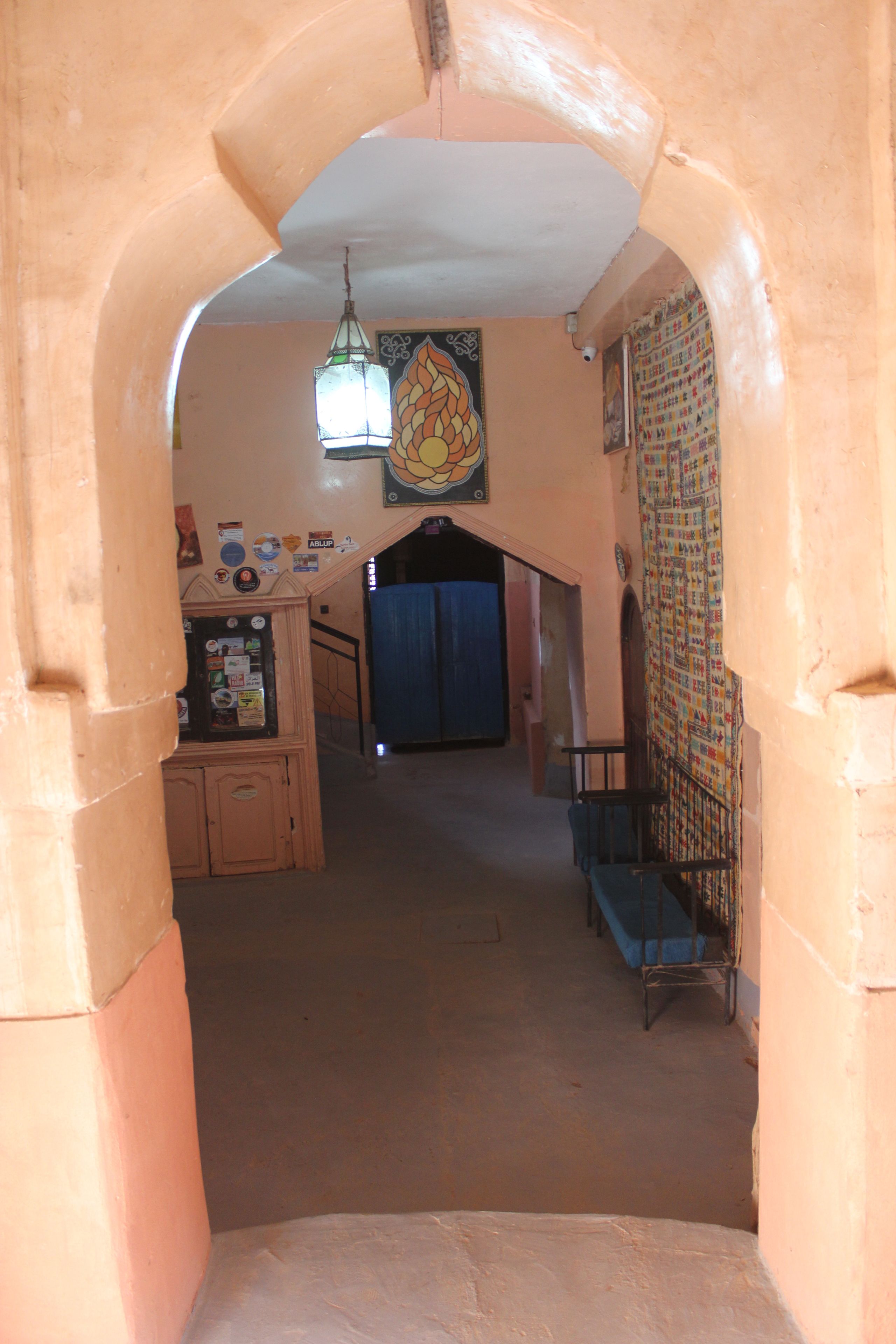 Un couloir étroit, avec une porte bleue au fond, un petit espace détente avec un banc en métal et un mur orné d’une tapisserie décorative.