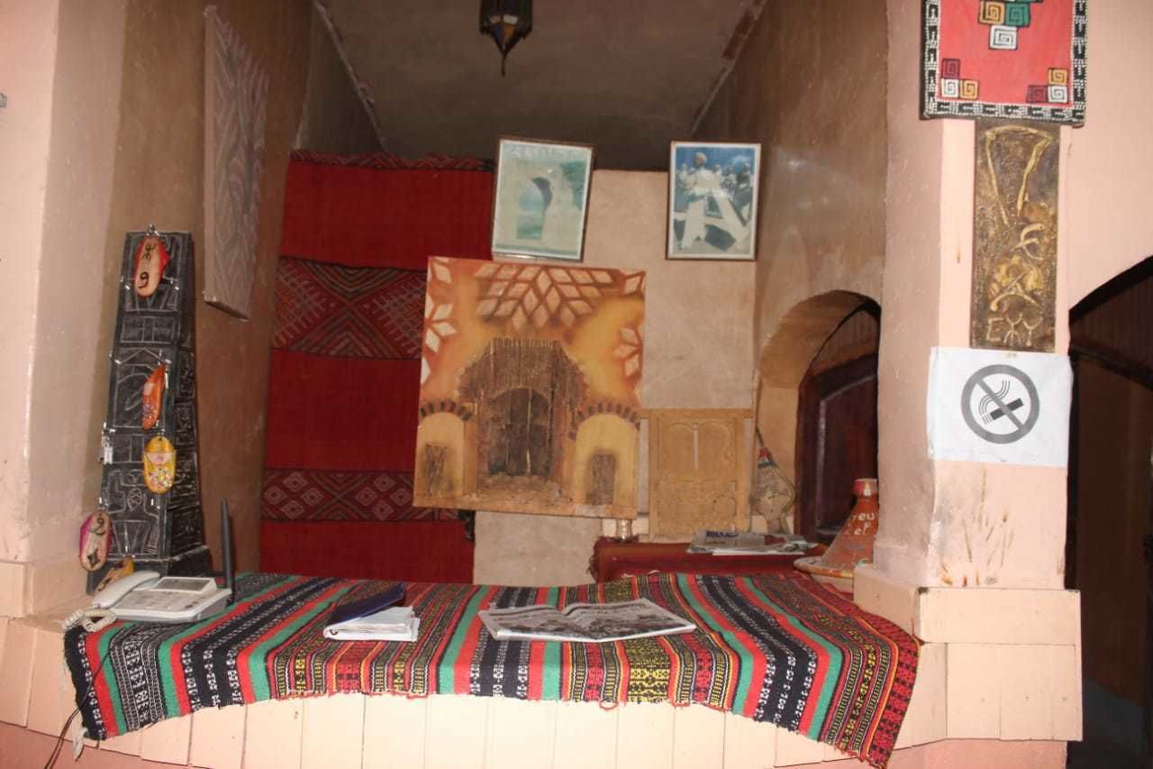 Una habitación con alfombra estampada, una cama con manta roja, un teléfono instalado en la pared y cuadros enmarcados.