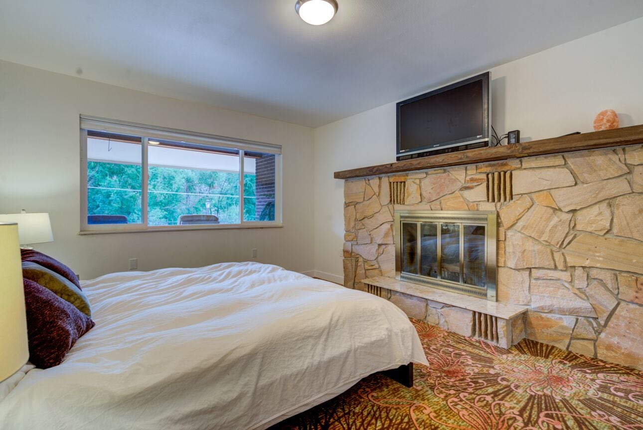 A bedroom with a stone fireplace, a bed with white bedding, a window with a view of trees, and a flat-screen TV mounted above the fireplace.