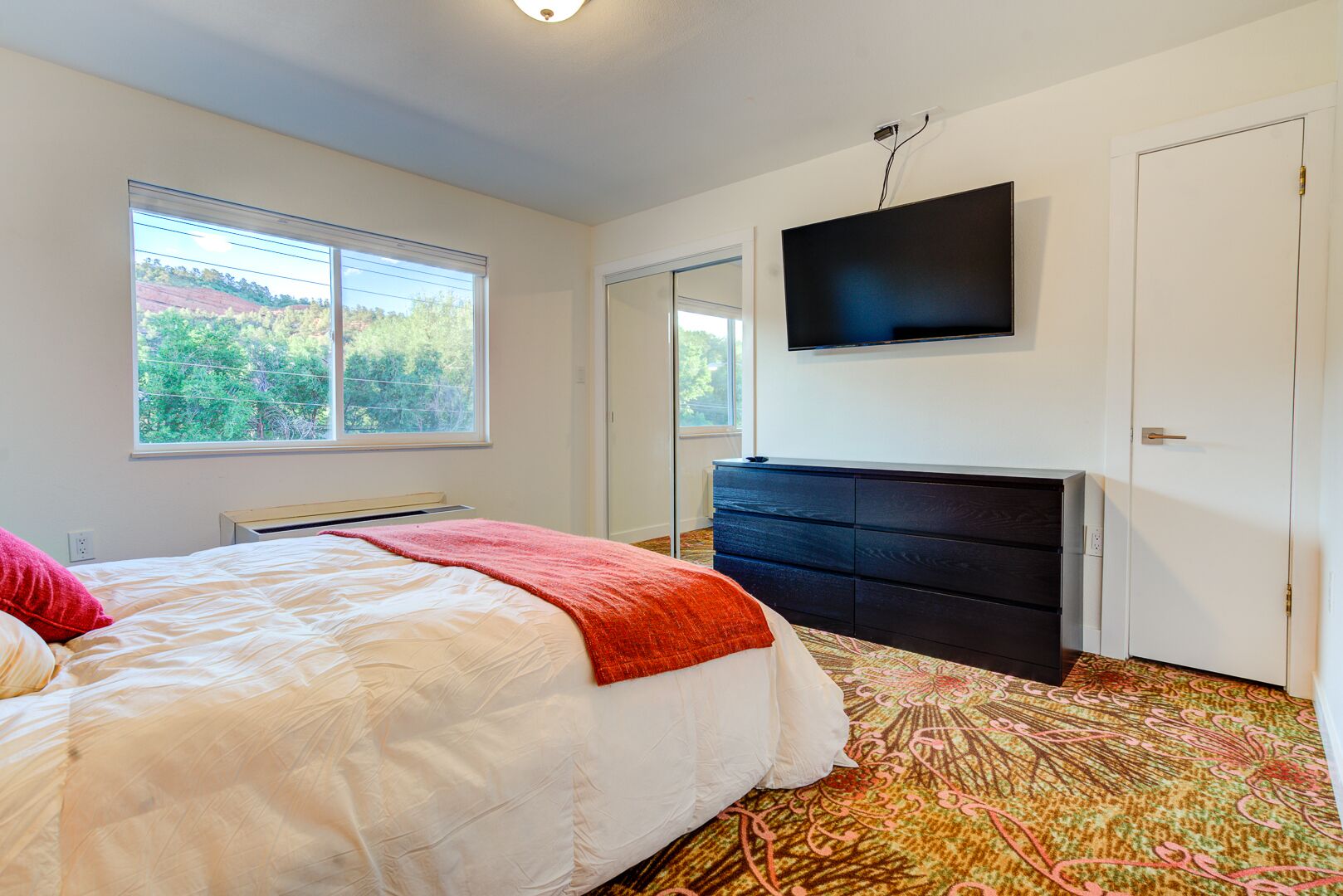 A bedroom with a bed, a dresser, a television mounted on the wall, and a window with a view of greenery.