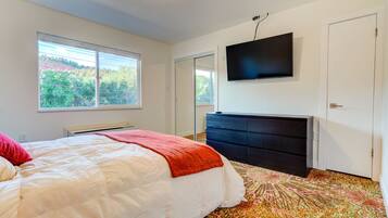 A bedroom with a bed, a dresser, a television mounted on the wall, and a window with a view of greenery.