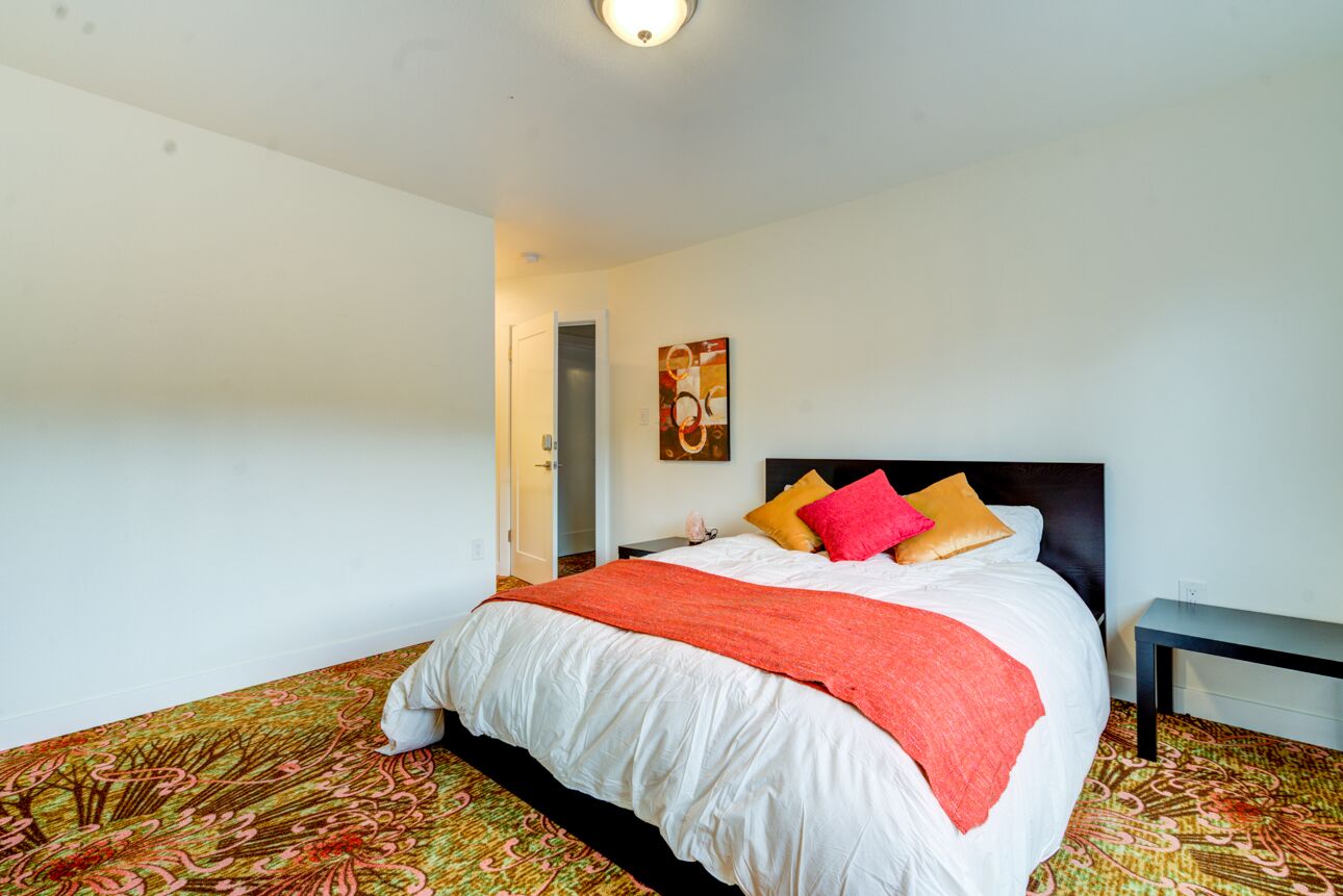 A bedroom with a large bed, a nightstand, a wall-mounted artwork, and a patterned carpet.