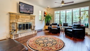 A living room with a stone fireplace, a flat-screen TV mounted above it, and a large window with a view of trees. There are two black leather sofas and a round rug on the wooden floor. - INNhale Resort and Spa (Manitou Springs)