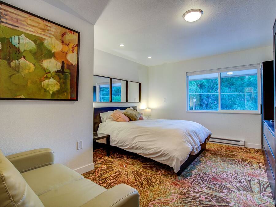 A bedroom with a bed, a sofa, a large painting, a window, and a mirror.