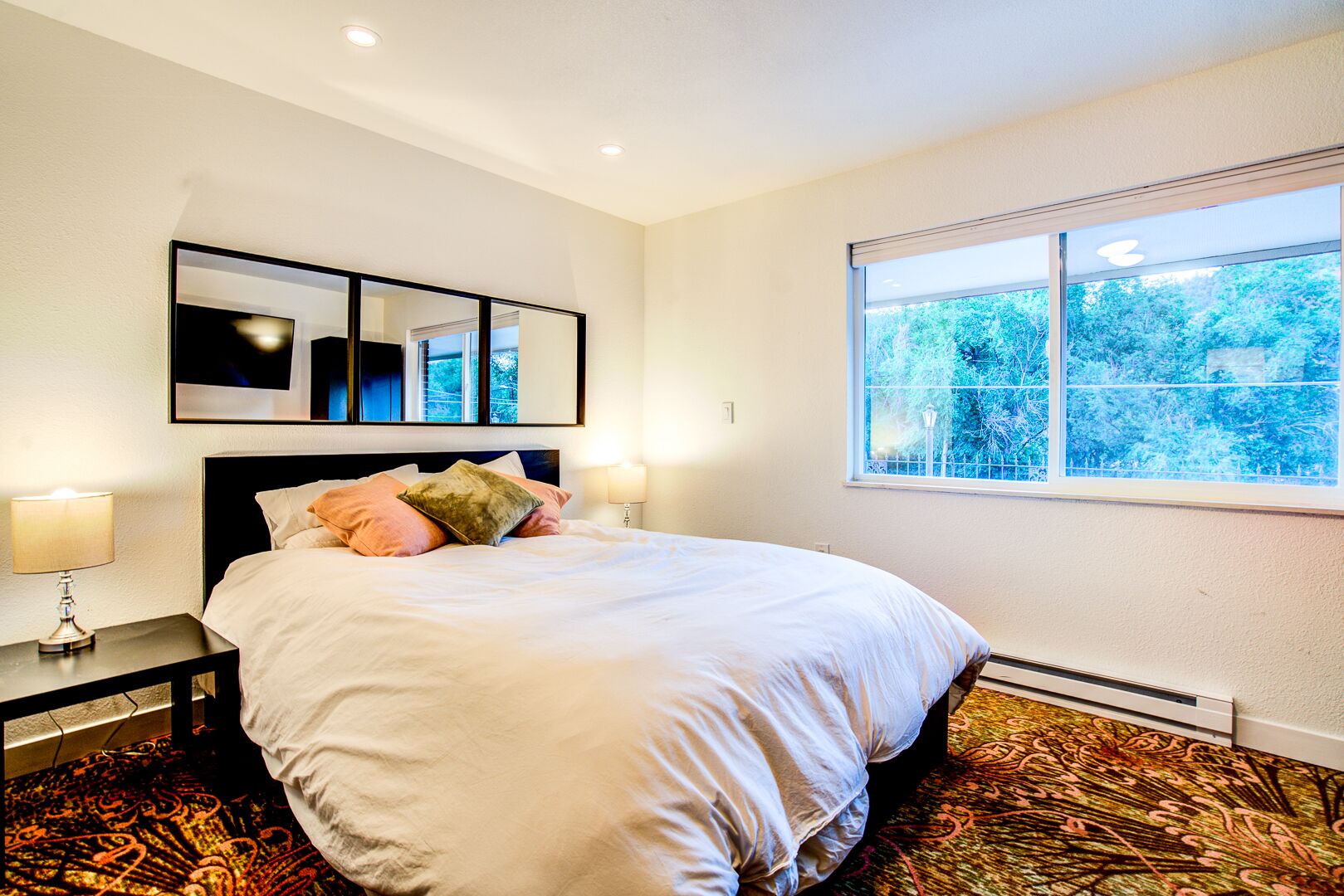 Une chambre à coucher bien aménagée, avec un lit, des lampes de chevet, une grande fenêtre donnant sur un paysage verdoyant et une œuvre d’art encadrée au-dessus du lit.