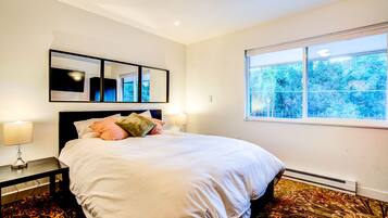 Une chambre à coucher bien aménagée, avec un lit, des lampes de chevet, une grande fenêtre donnant sur un paysage verdoyant et une œuvre d’art encadrée au-dessus du lit.