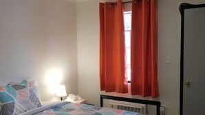 A bedroom with a bed, a nightstand, a window with orange curtains, and a radiator. - Spruce Pine Inn (Spruce Pine)