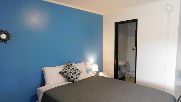A hotel room with a blue wall, a bed with a gray quilt, white pillows, a nightstand with a lamp, and a bathroom visible through an open door. - Spruce Pine Inn (Spruce Pine)