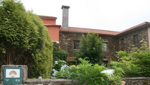 A stone building with a red-tiled roof, a chimney, and a green sign with a logo. - Casa Pousadoira (Mino)