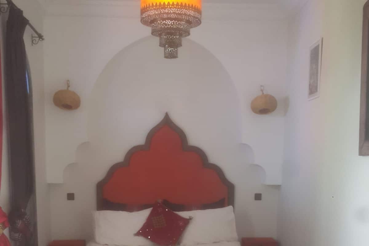 A bedroom with a bed, a round wooden table, a green sofa, a hanging lamp, and a patterned rug.
