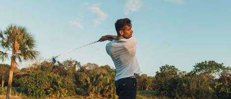 A person playing golf on a well-maintained course with trees and a clear sky in the background.