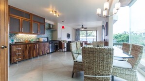 A modern kitchen with wooden cabinets, a dining area with wicker chairs, and a large window offering a view of greenery. - Mimosa Flat by Realty Group (Playa del Carmen)