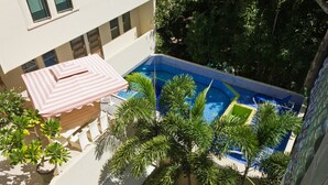 En pool på taket med ett randigt parasoll, omgiven av frodig grönska och en vit byggnad.