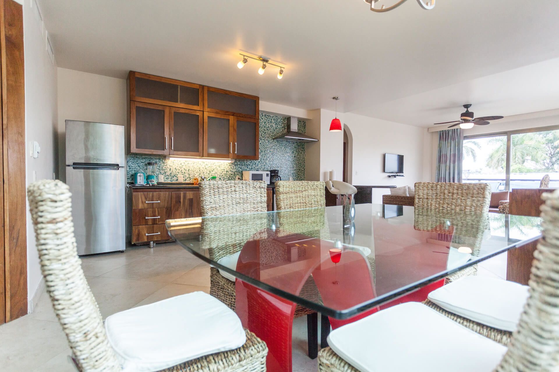 Una cocina moderna con armarios de madera, un refrigerador de acero inoxidable y un comedor con mesa de vidrio y sillas de mimbre.