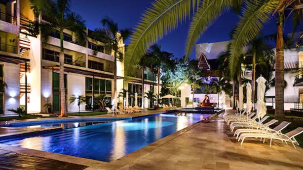Zona junto a la piscina con sillones, palmeras y un edificio de varios pisos con balcones.