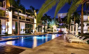Un espace aménagé au bord de la piscine, avec des chaises longues, des palmiers et un bâtiment à plusieurs étages doté de balcons.