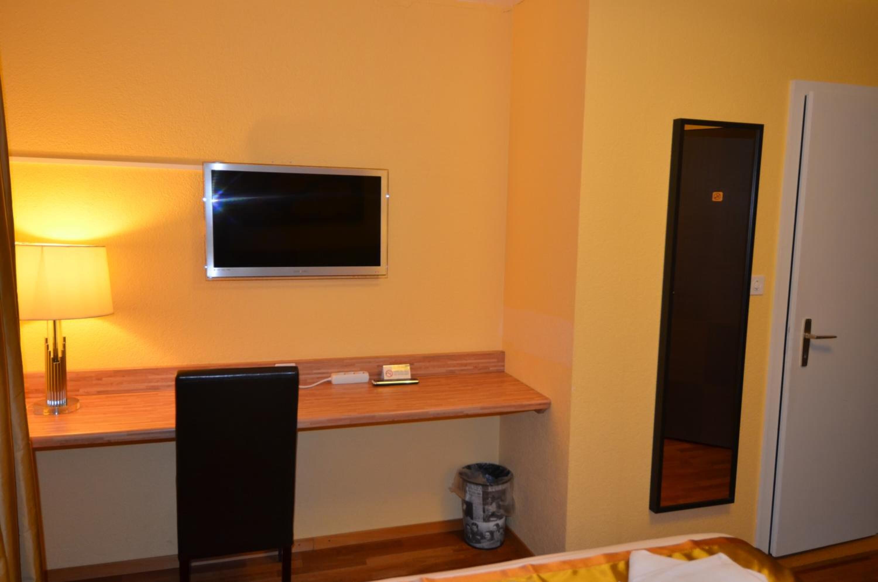 A hotel room with a wooden desk, a television mounted on the wall, a chair, a small trash bin, and a door.