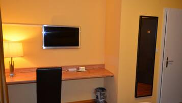 A hotel room with a wooden desk, a television mounted on the wall, a chair, a small trash bin, and a door.