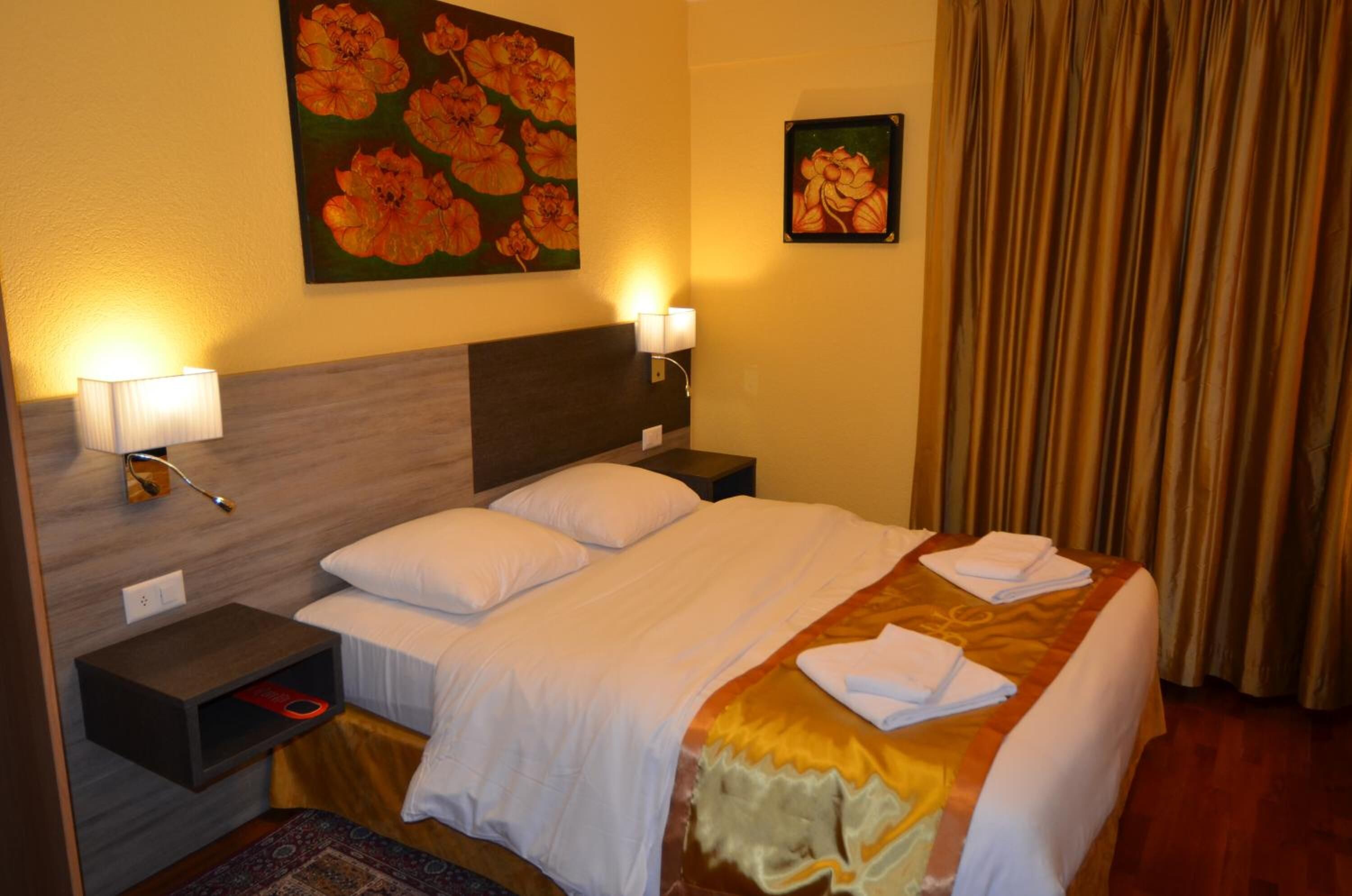 A hotel room with a bed, two bedside lamps, a nightstand, a painting of flowers, and a curtain.