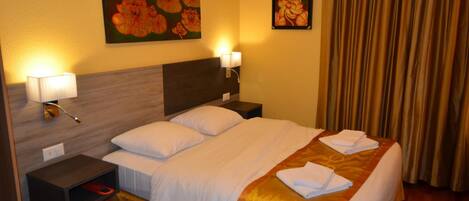 A hotel room with a bed, two bedside lamps, a nightstand, a painting of flowers, and a curtain.