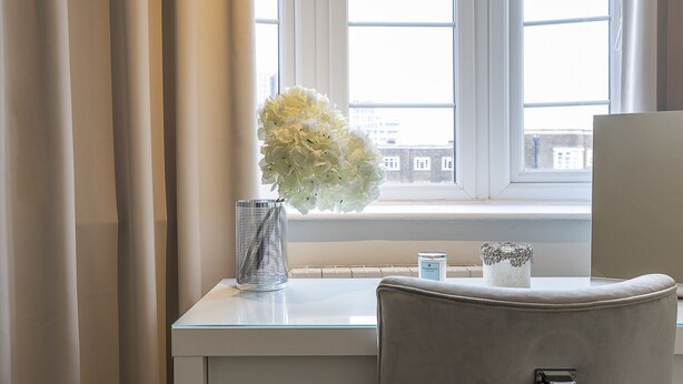 Une pièce avec un bureau, un vase de fleurs, une bougie et une vue sur des immeubles par la fenêtre.