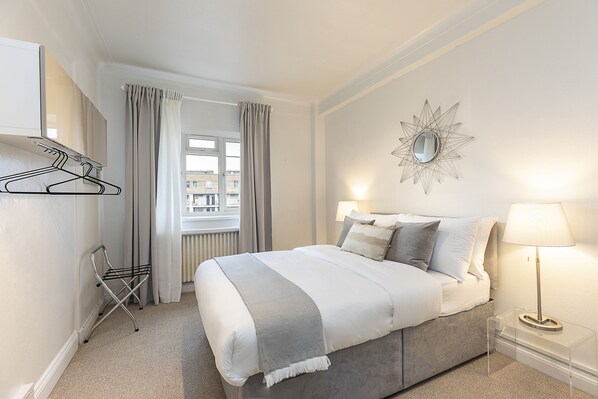 Une chambre à coucher bien rangée, avec un lit, des lampes de chevet, un porte-manteau suspendu et une fenêtre avec des rideaux.