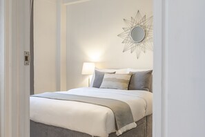 A neatly made bed with a grey throw blanket, a wall-mounted mirror, and a bedside lamp. - Grand Swiss Cottage Apartment (London)