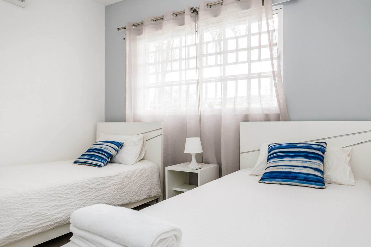 Une chambre d’hôtel avec deux lits, une table de chevet avec une lampe et une fenêtre avec des rideaux légers.