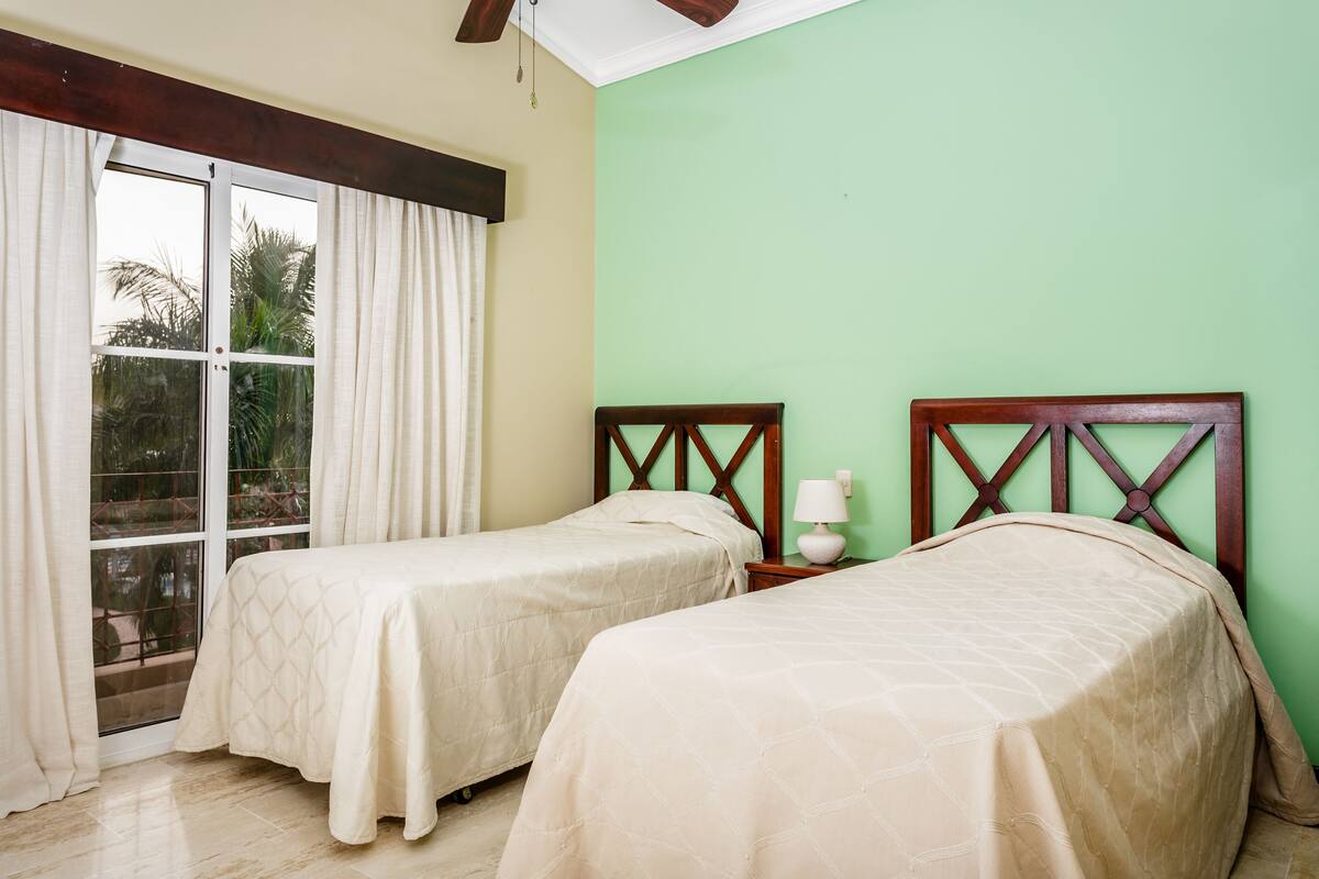 Une chambre avec deux lits, une fenêtre avec des rideaux, une table de chevet avec une lampe et un mur vert.