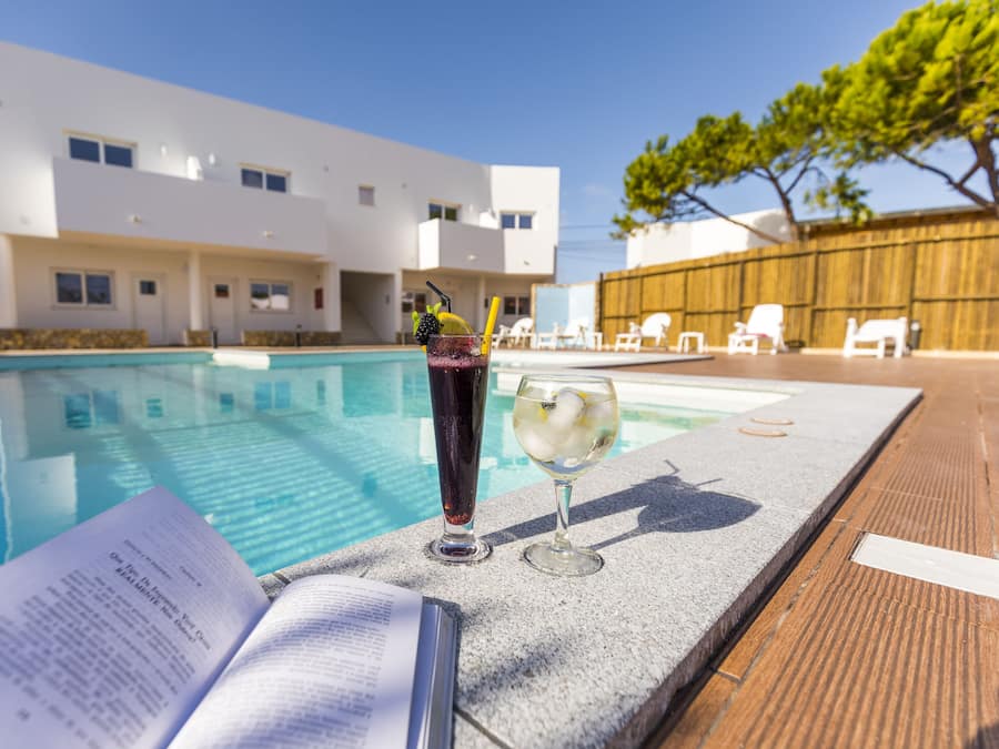 Escena junto a la piscina con un libro, dos bebidas y sillones de descanso.