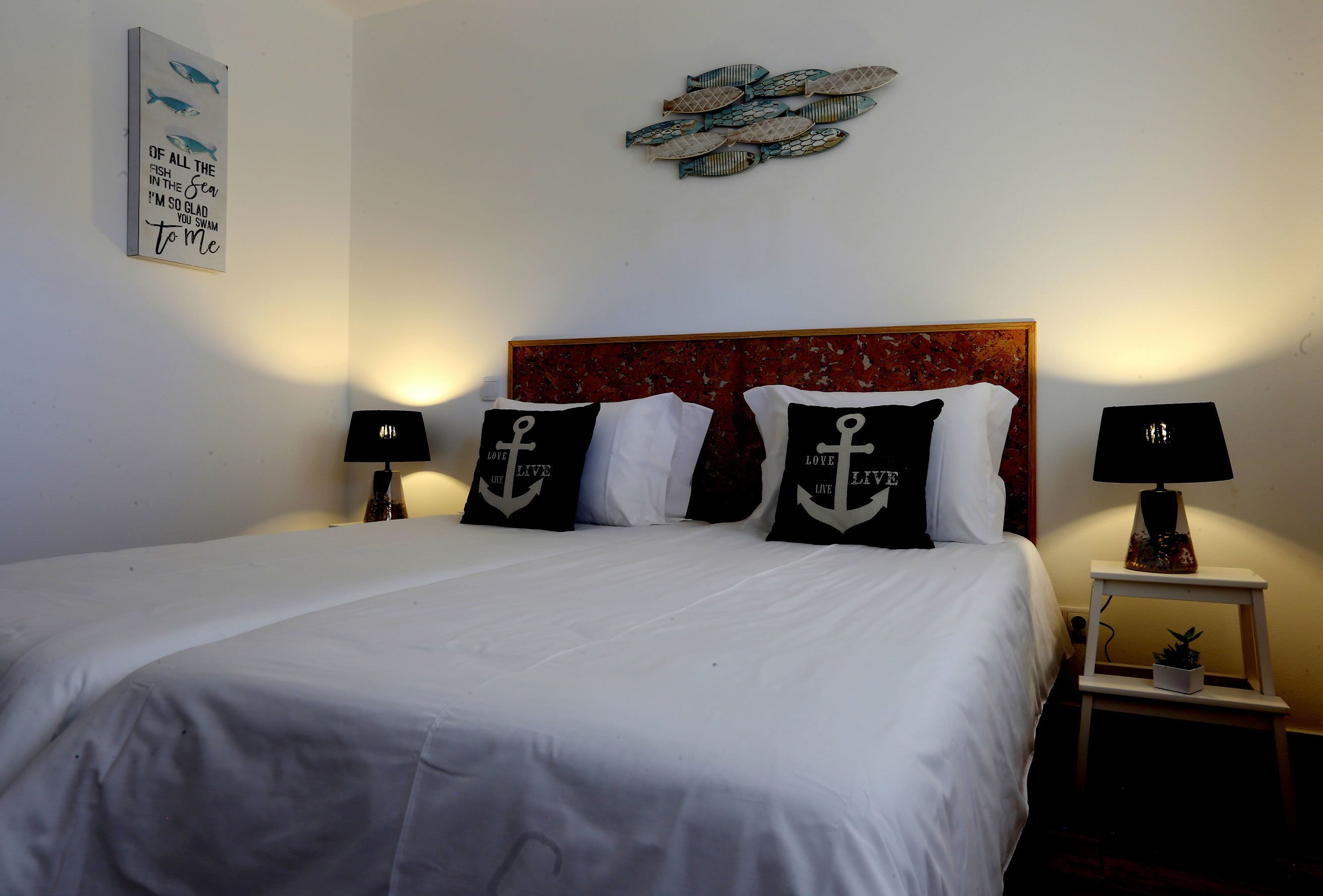 Ein ordentlich bezogenes Bett mit weißer Bettwäsche und schwarzen Kissen mit Anker-Motiven, ein roter Kopfteil und ein Wandbild mit Fischen.